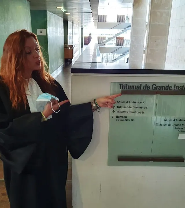 En robe noire, désignant du doigt un panneau « Tribunal de Grande Instance »
