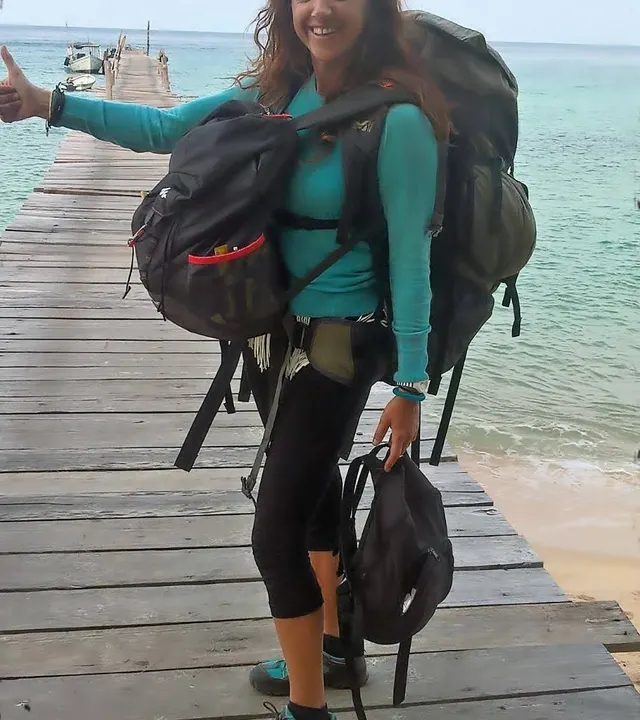 Sac à dos sur le dos, pouce levé, sur un ponton en bois au bord d’une mer turquoise